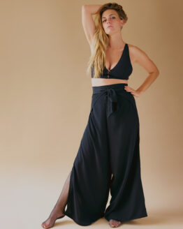 Femme debout se tenant les cheveux porte un petit top noir et un pantalon fendu noir avec des résilles