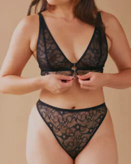 Femme debout portant un ensemble en lingerie noire rattache son soutien gorge à l'aide de petits crochets métallique